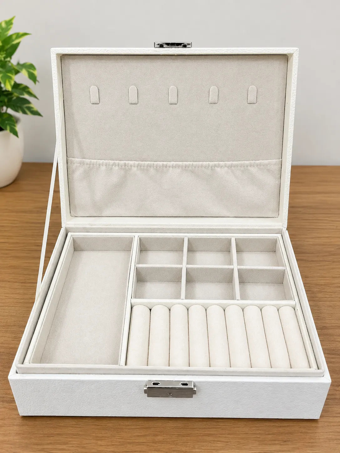 An open white jewelry organizer box with a textured exterior and cream-colored velvet interior, featuring compartments, ring rolls, and a hanger section on the lid, displayed on a wooden tabletop.