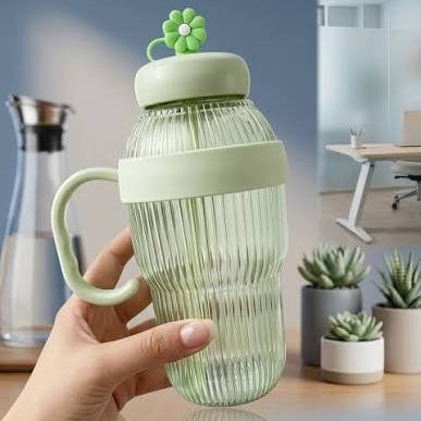 A hand holds a ribbed translucent light green water bottle with a matching green handle, lid, and flower-shaped lid topper, set against an office-like background with a desk and small potted succulents.