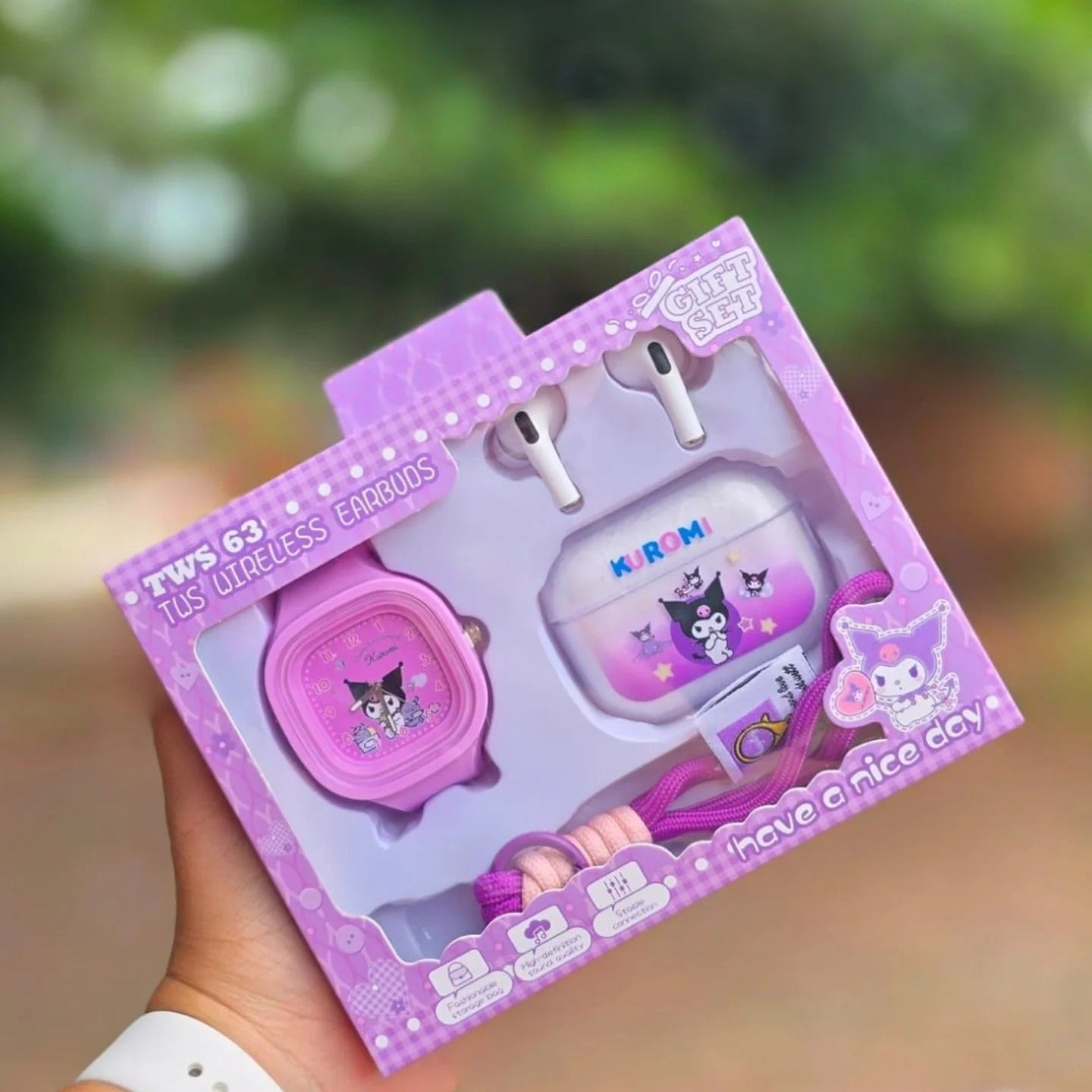 A hand holding a pink and purple toy set in retail packaging, featuring a cartoon watch, earbud case, and accessories, with a blurred outdoor background.