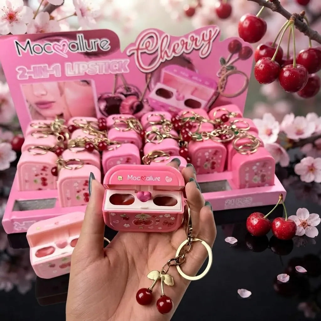 A hand holds a pink cherry-themed lipstick compact in front of a display of matching lipsticks in a box labeled 'Mocvalure Cherry 2-in-1 Lipstick'. The compact has cherry illustrations and a gold keychain with red cherries.