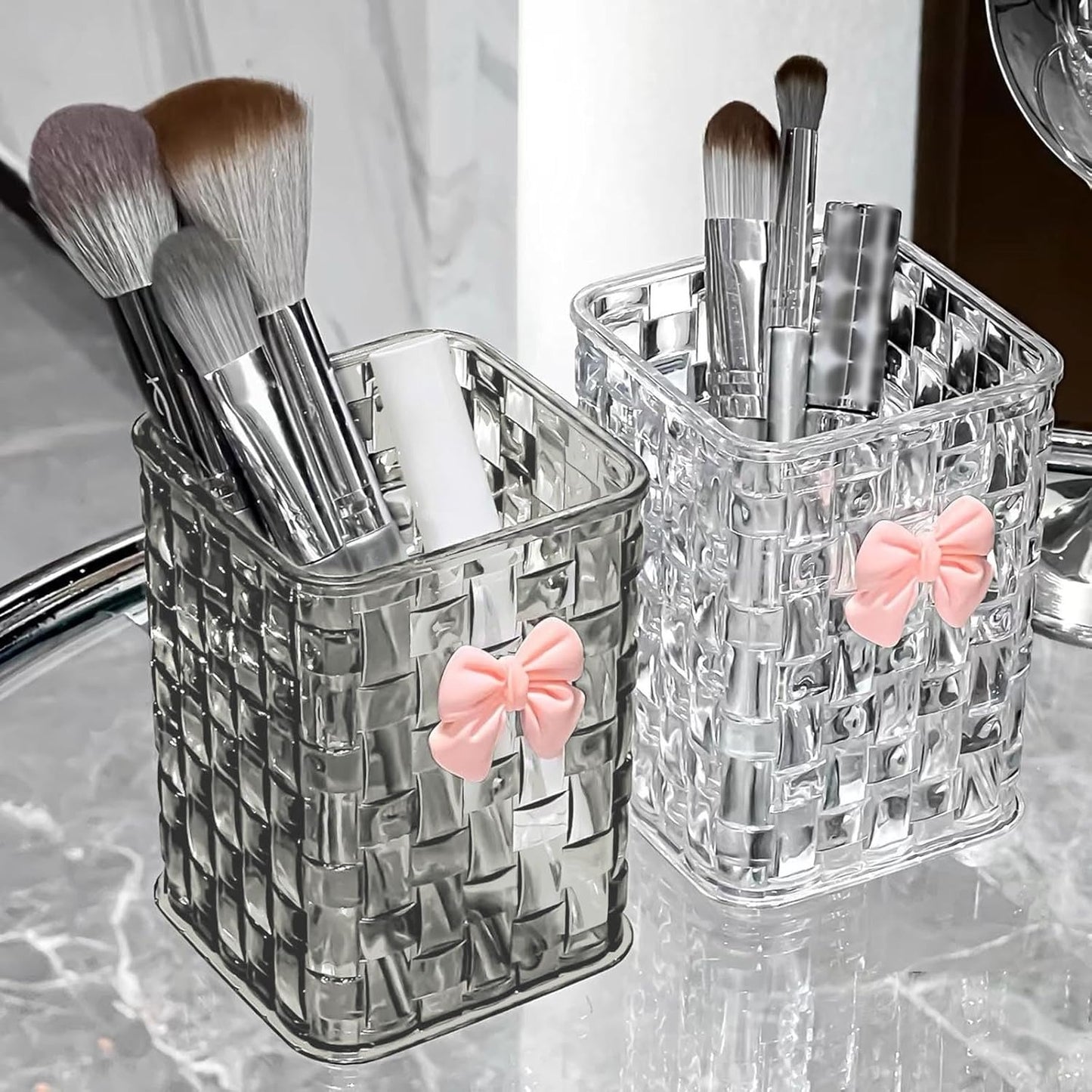 Two clear textured plastic makeup brush holders with pink bow decorations, shown on a marble countertop with makeup brushes inside.