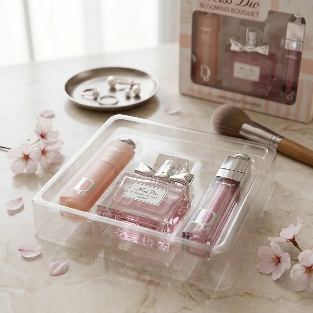 A clear plastic organizer tray holding a pink perfume bottle, a pink lip product, and a pink cylindrical item, styled on a marble surface with flowers, a makeup brush, rings, and a boxed set in the background.