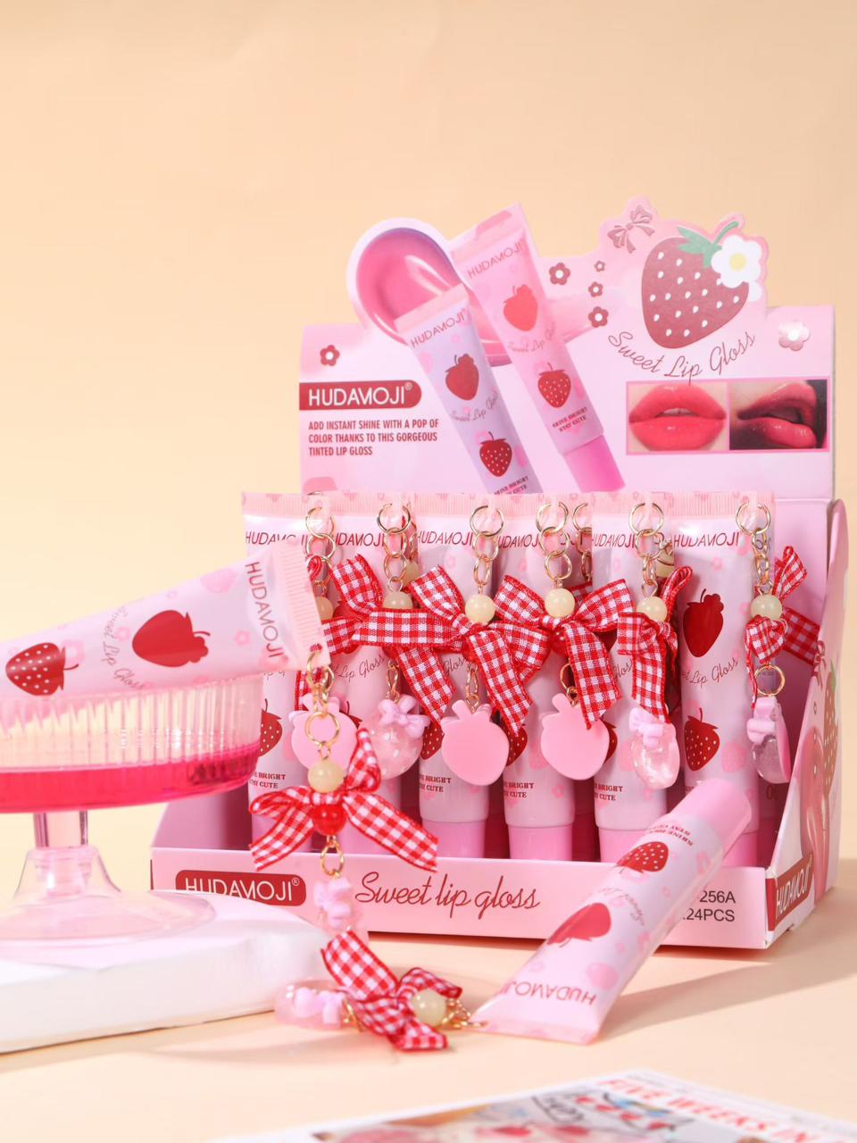 A pink and strawberry-themed HUDA MOJI Sweet Lip Gloss display set, featuring multiple lip gloss tubes, a lip brush, and decorative pink accessories with red gingham bows and strawberry illustrations, arranged on a light beige background.