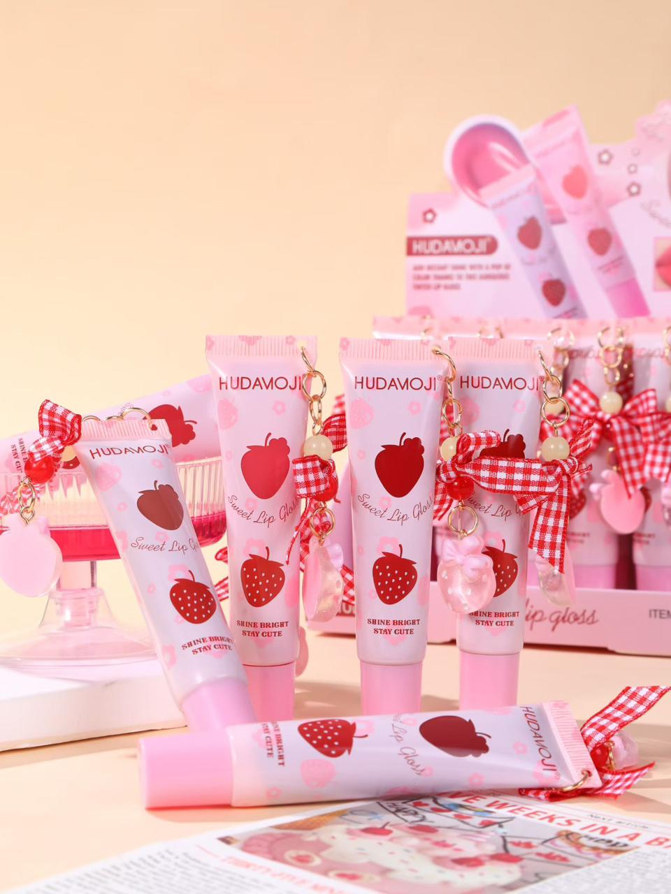 Pink strawberry-themed cosmetic tubes and boxes with red apple prints, gingham ribbon bows, and gold charms, arranged on a light beige surface against a soft peach background.