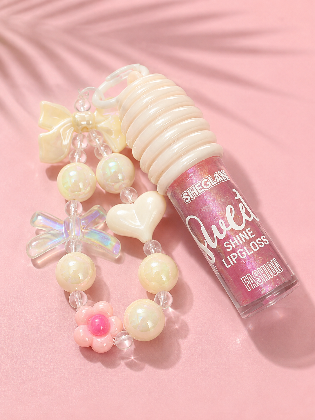 Sheglan Sweet Shine Lipgloss in a pink tube with a ribbed white cap, styled with a decorative beaded charm strap on a pink background.