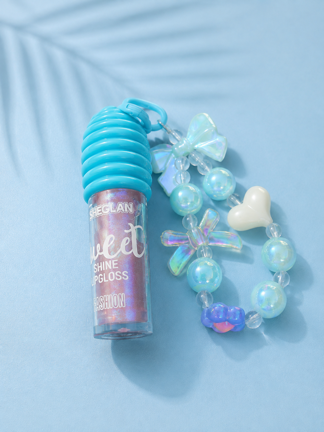 A tube of Meglan Sweet Shine Lipgloss in a pink holographic shade, styled next to a matching iridescent bead charm bracelet with a turquoise ribbed cap on a light blue background.