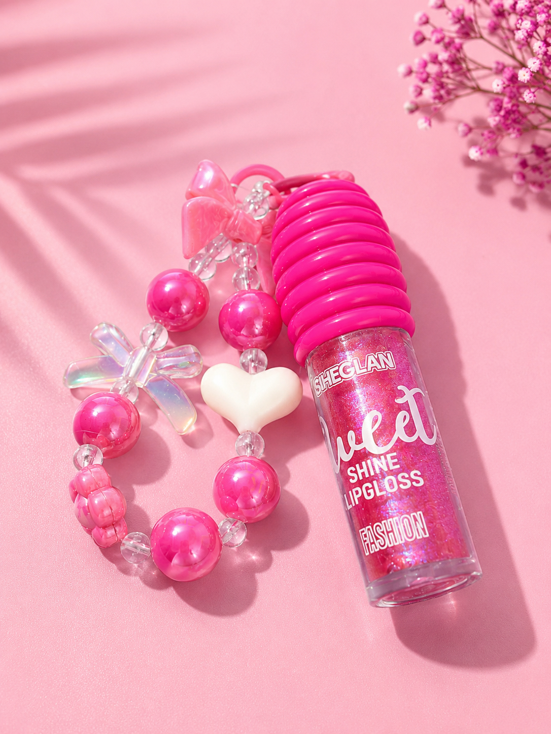A pink lip gloss tube labeled 'SIEGLAN Sweet Shine Lipgloss' placed next to a pink beaded charm bracelet with a white heart, on a pink background with decorative dried flowers and soft shadows.