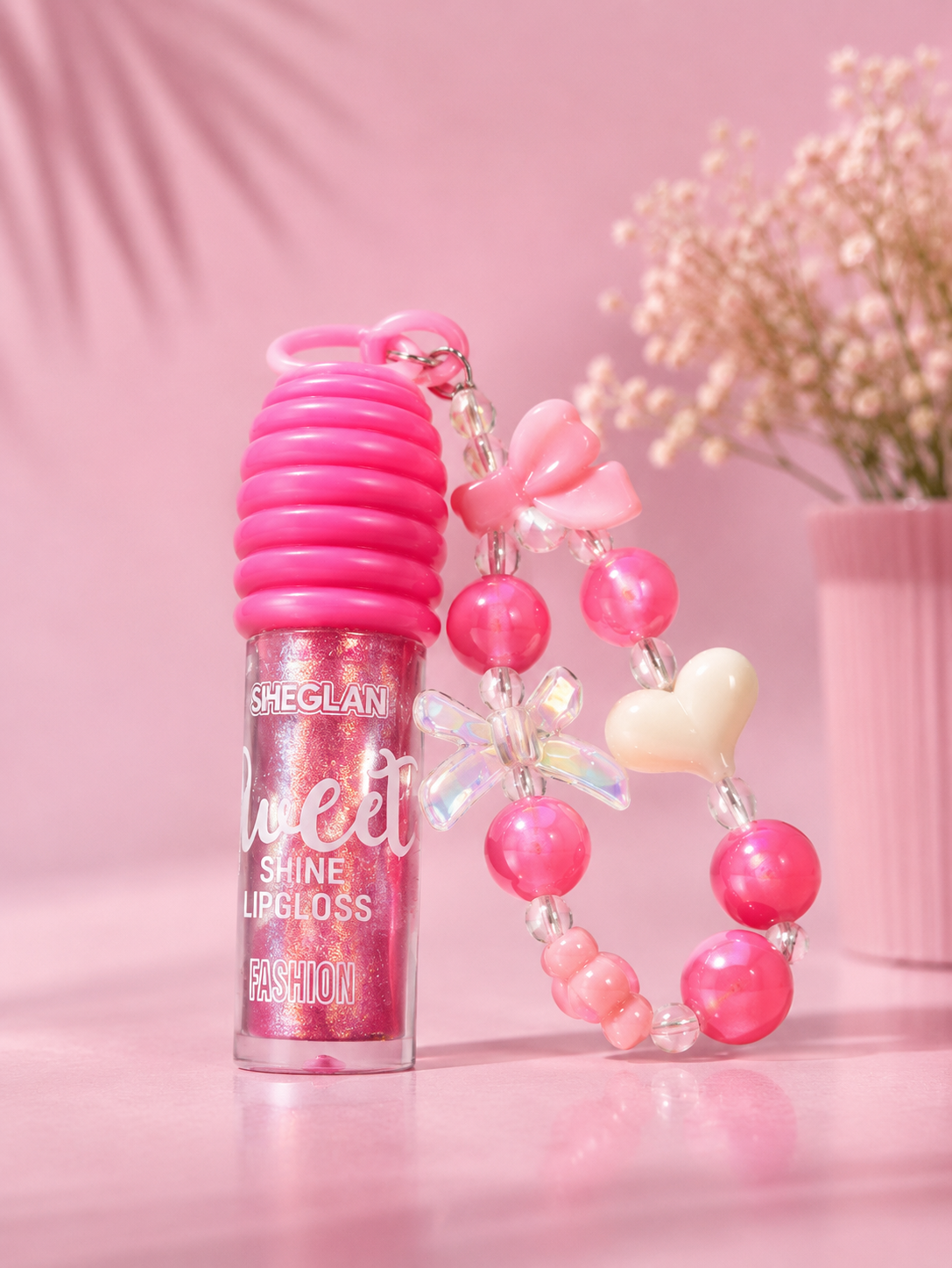 Sheglani Sweet Shine Lipgloss in a pink bottle with a matching pink ribbed cap, shown with a pink beaded wristlet strap featuring bows, pearls, and a heart charm on a pink background.