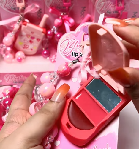 A close-up of a person holding a pink heart-shaped lip color compact with an open translucent lid, surrounded by matching pink lip glosses and decorative accessories.