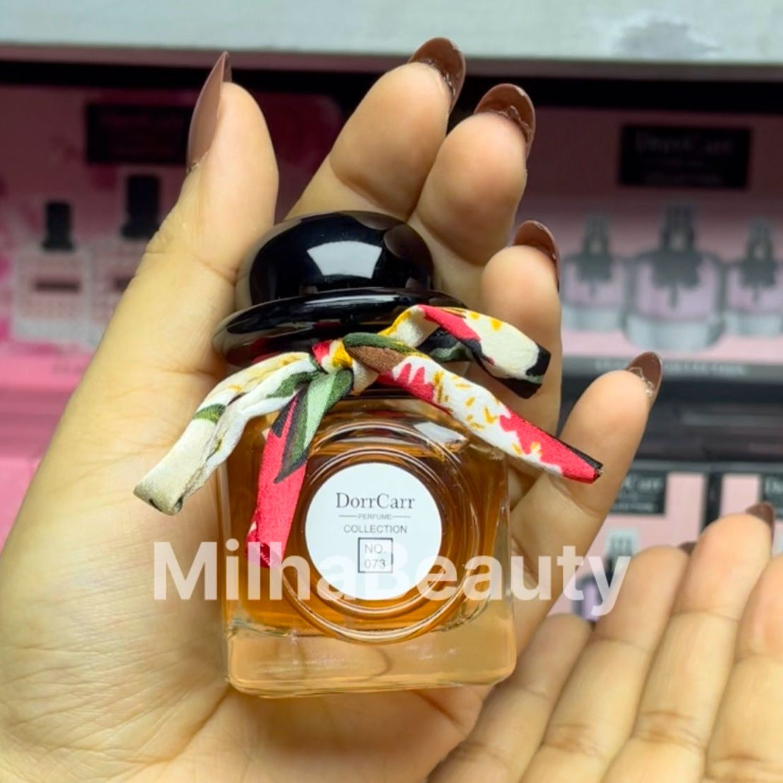 Hand holding a small bottle of Dorrcarr perfume with a colorful ribbon, against a blurred background of more perfume bottles.