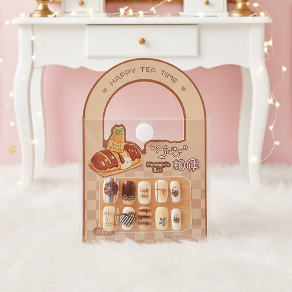 A 10-piece press-on nail set in a clear plastic blister pack with a peach-colored arch header card, placed on top of a black laptop keyboard. The nails feature various cute tea-themed illustrations like a tea bag, a cup of black tea, a matcha leaf, and a chocolate bar.