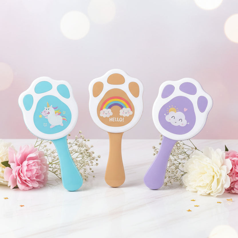 Three packaged pet grooming brushes with paw-shaped heads are displayed on a colorful tropical-patterned fabric. The left brush is light blue with a unicorn design, the middle is tan with a rainbow design, and the right is pink with a cloud design.