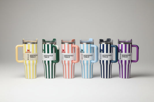 Six insulated handled tumblers with vertical striped patterns in different colors, displayed on a glass shelf in front of cardboard boxes.