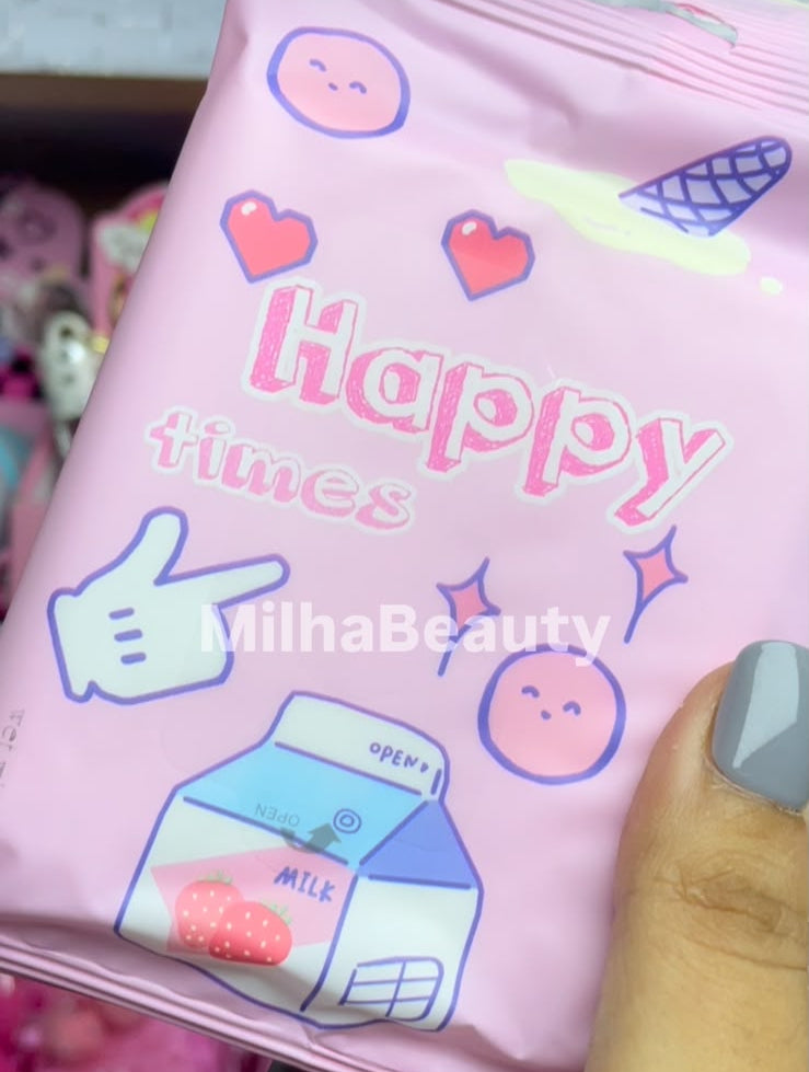 A hand holding a pink packet of MilhaBeauty Happy Times lip balm, with a close-up of the product packaging featuring cartoon illustrations and text.