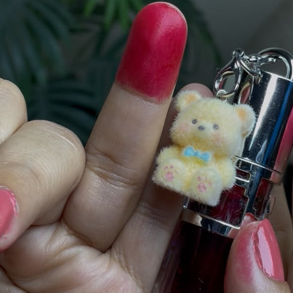 A close-up of a hand holding a small tube of dark red nail polish or lip gloss with a plush teddy bear charm attached. The hand has bright pink nails, and the background shows a soft-focus indoor setting with green.leaves.
