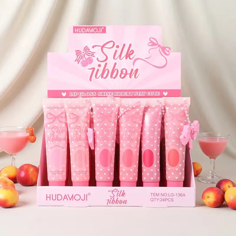 A display box of Hudamoji Silk Ribbon lip balm tubes in shades of pink, decorated with hearts and bows, styled with pink drinks and peaches on a draped cream fabric background.