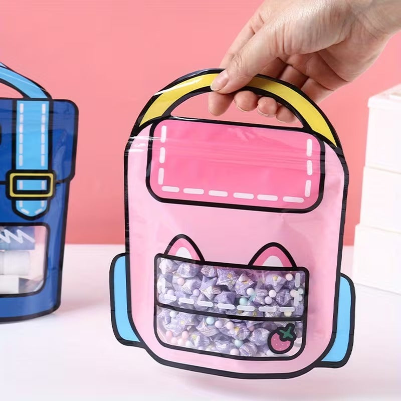A hand holds a pink cat-shaped clear plastic snack bag with a yellow handle and blue accents, with a blue bag in the background against a pink backdrop.