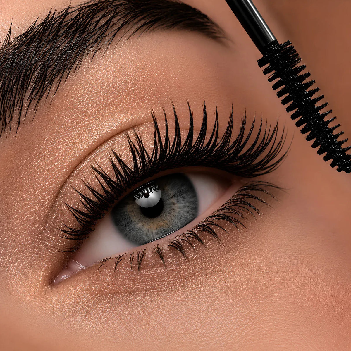 Close-up of a person's eye with long, curled black eyelashes while a black mascara wand applies product near the upper lash line.