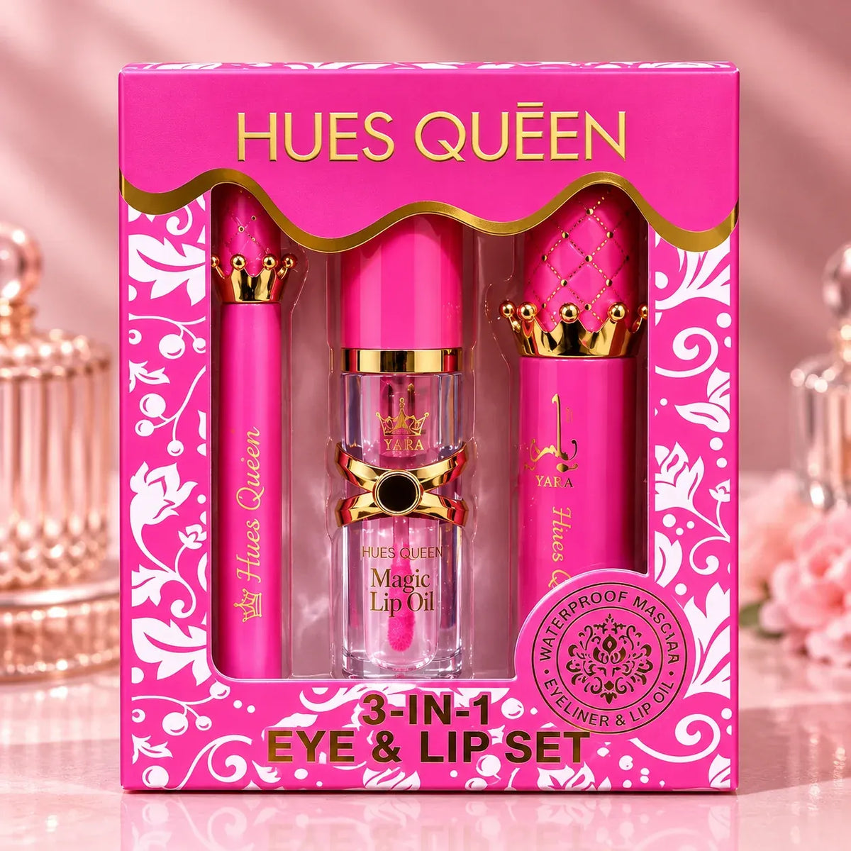 Hues Queen 3-in-1 Eye & Lip Set in a bright pink gift box with gold lettering and decorative swirls. The set includes a pink lip oil bottle in the center and two matching pink lip products with gold crown caps on either side, shown against a soft pink background.