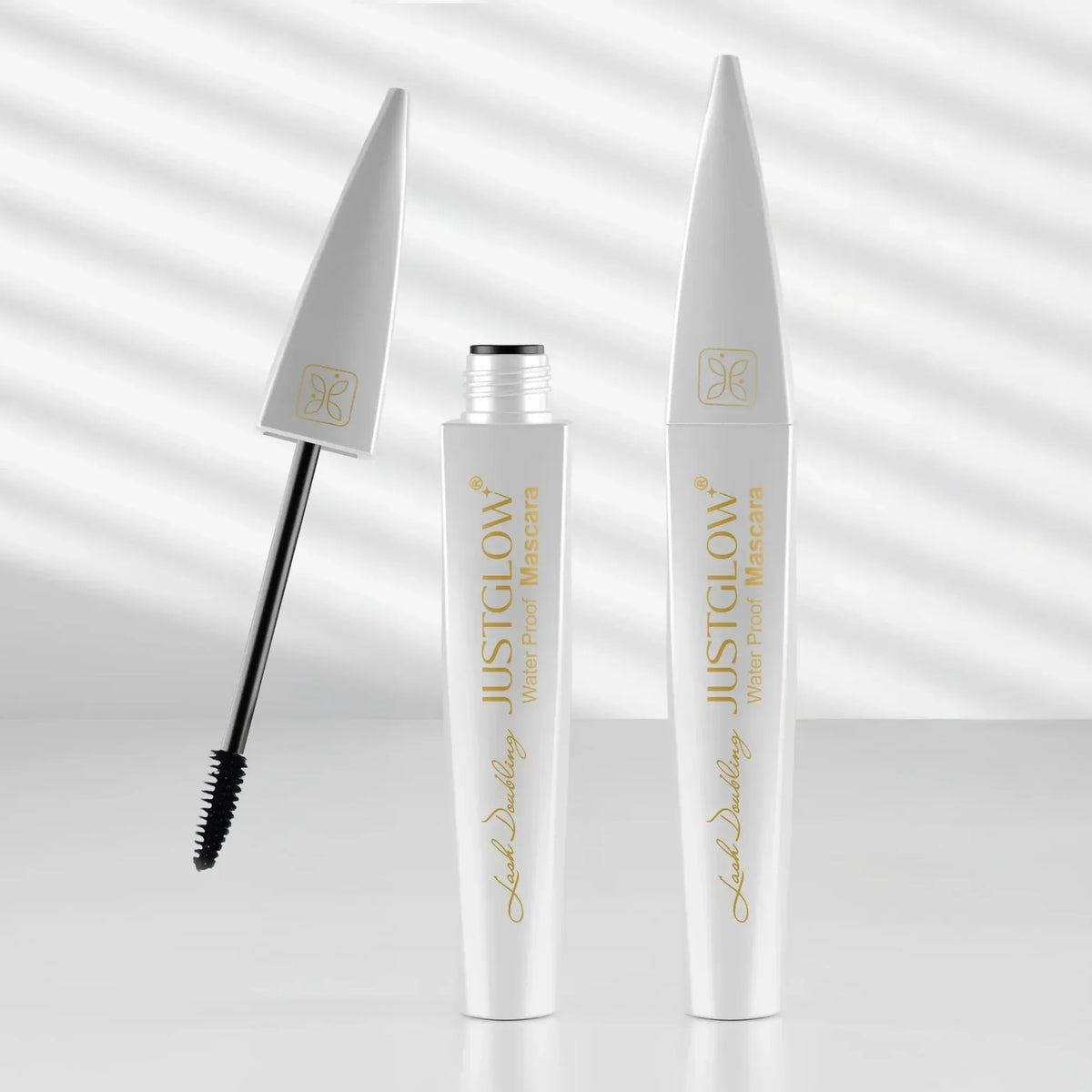 White mascara tubes with gold branding and a black mascara wand, shown on a clean white background with soft diagonal shadows. One tube is open with the wand beside it, and one tube stands closed.