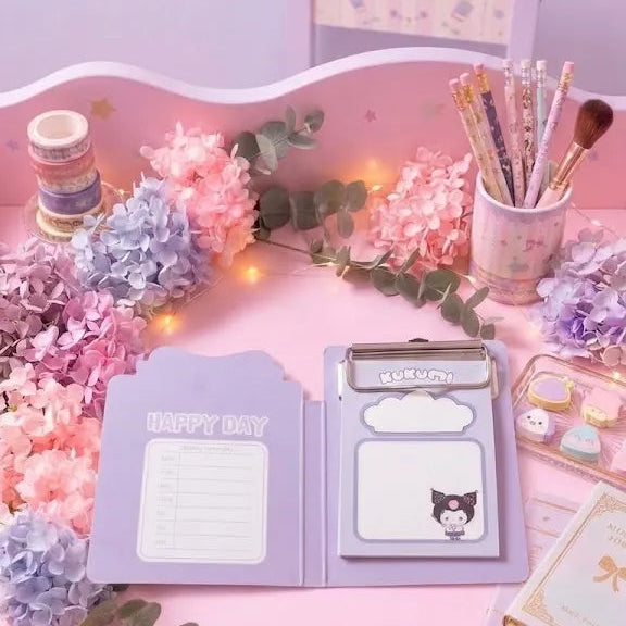 A pastel pink desk setup with a lavender clipboard notepad and sticker sheet, surrounded by pink and purple flowers, eucalyptus stems, a cup of pens, and small stationery items on a matching pink desk.