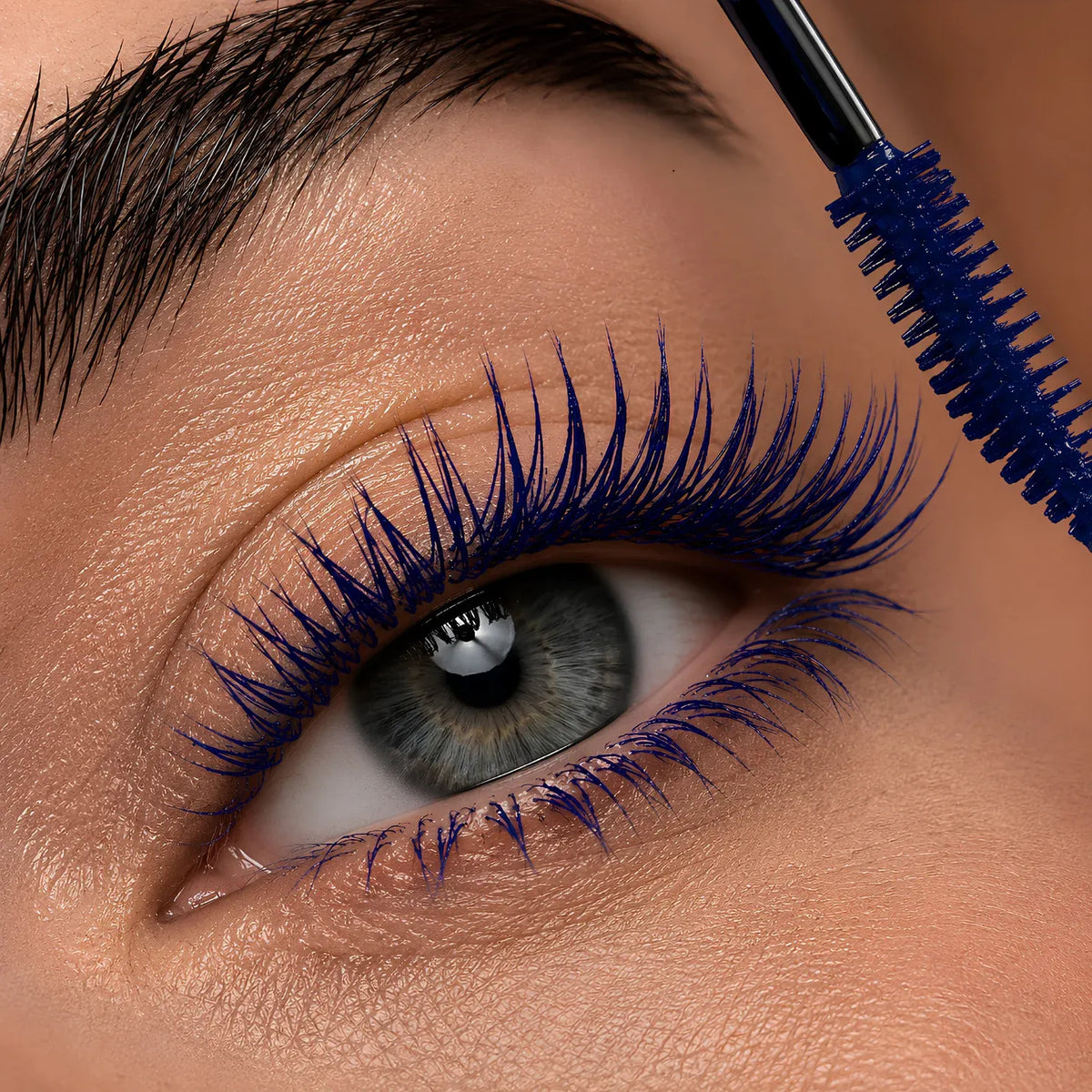 Close-up of a person's eye with long, dark blue mascara lashes being coated by a blue mascara wand.
