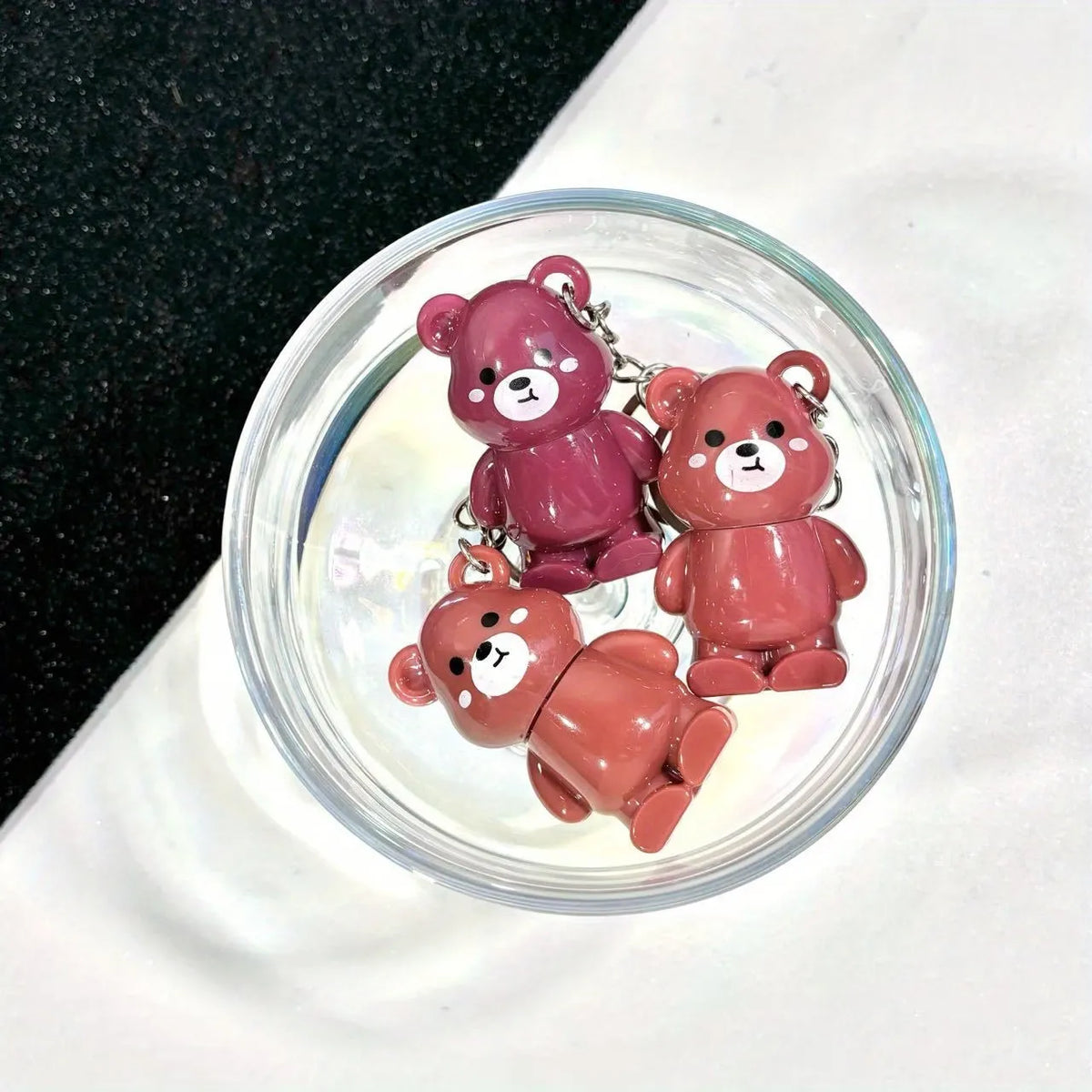 Three small bear-shaped keychains in pink, mauve, and terracotta red, arranged inside a clear round glass dish on a white surface with a black textured area in the background.