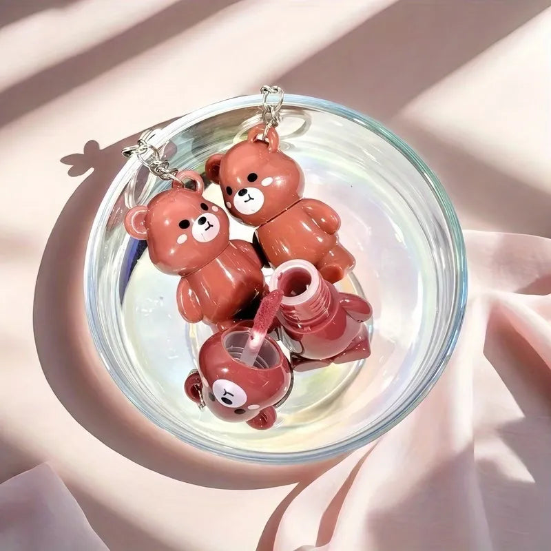 Three small bear-shaped lip balm tubes in a glossy terracotta pink color, arranged inside a clear glass dish on draped pink fabric with soft sunlight shadows.