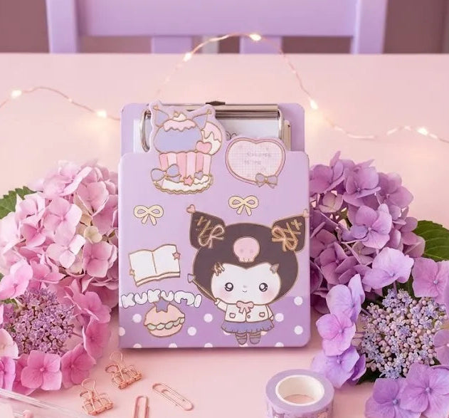 A pastel purple clipboard with cute cartoon character stickers and the word 'KUMUSHI' displayed on a pink table, surrounded by pink and purple hydrangeas, stationery, and fairy lights in a styled room setting.