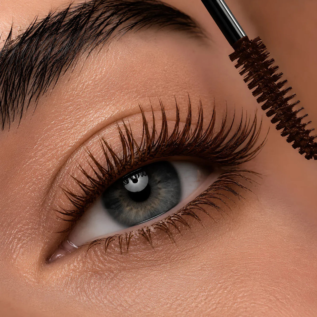Close-up of a person's eye with long, dark eyelashes while a spoolie wand applies mascara. The eyebrow and surrounding skin are also visible in this tight beauty shot.