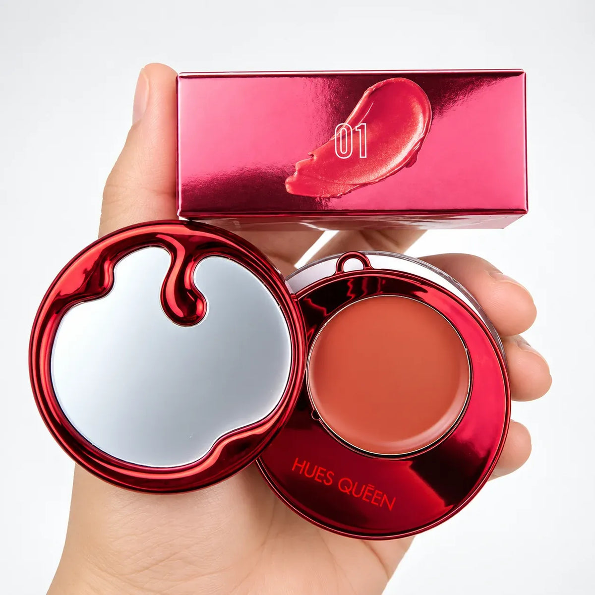 A hand holding a red makeup compact with a mirror lid, an open round compact showing a peachy coral cream product, and a matching metallic red box labeled '01' on a white background.