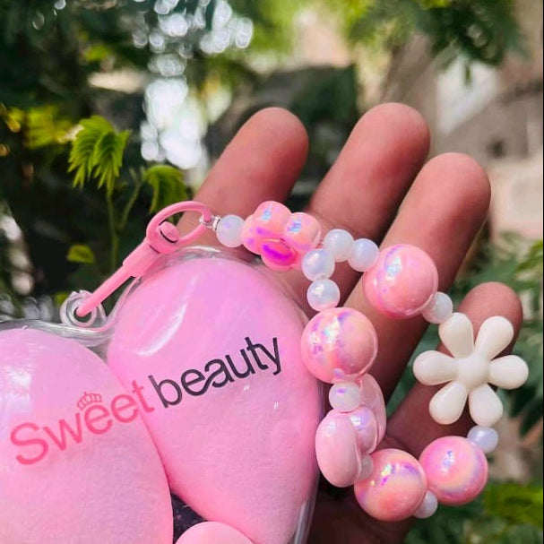 A hand holding a clear heart-shaped makeup blender case containing three pink sponge blenders, with a pink charm bracelet attached. The background is a blurred green outdoor foliage.