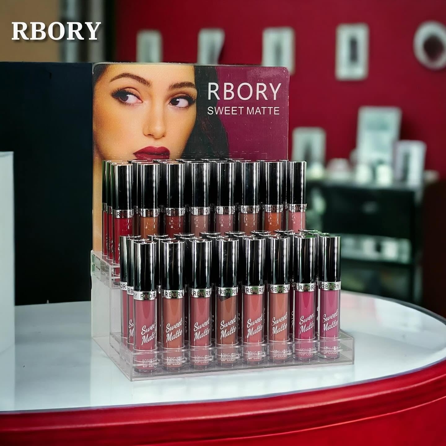 A clear acrylic display stand holding multiple tubes of RBORY Sweet Matte liquid lip glosses in various nude and pink shades, with the brand card behind them on a red and black surface.