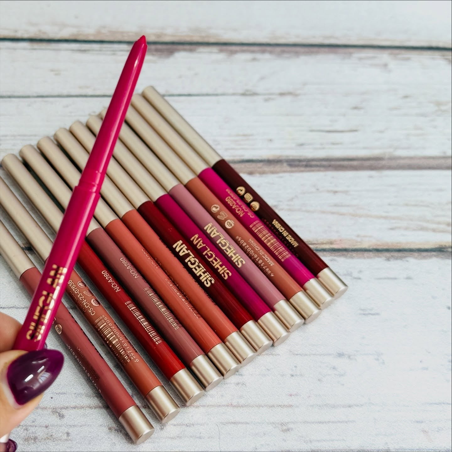 A hand with dark purple nail polish holds a bright pink lip pencil over a fan of SWIGELAN lipsticks in various shades, set against a white-washed wooden background.
