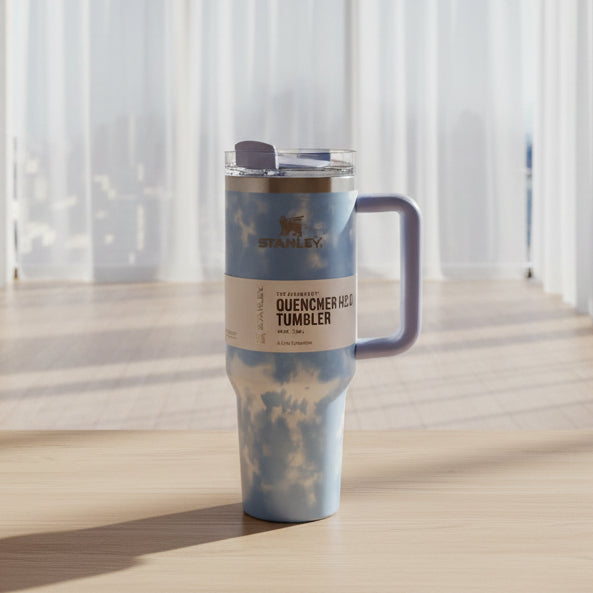 A hand holding a Stanley Quincher 22 in. tumbler with a sky-blue and white cloud pattern, a light blue handle, and a clear lid, set against a home background.