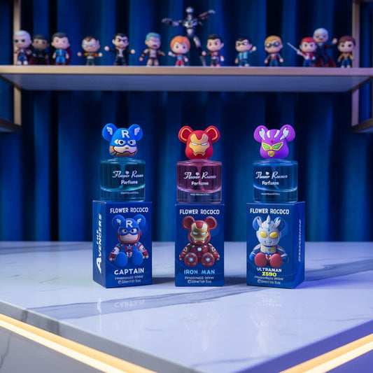 Three Flower Rococo perfume bottles with cartoon character caps and matching blue boxes are displayed on a wooden surface against a wood panel background. The characters shown are Captain America, Iron Man, and Ultraman.