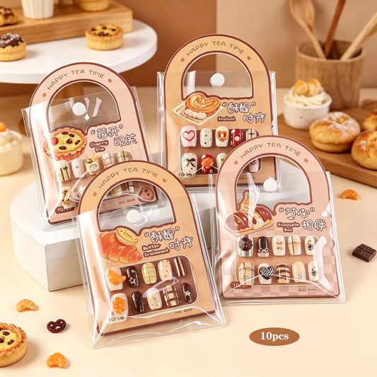 Four packaged sets of decorative press-on nails arranged on a beige tabletop with pastry props in the background. The packages are arched and labeled 'HAPPY TEA TIME' with illustrations of cakes, pastries, and candy-like nail designs.