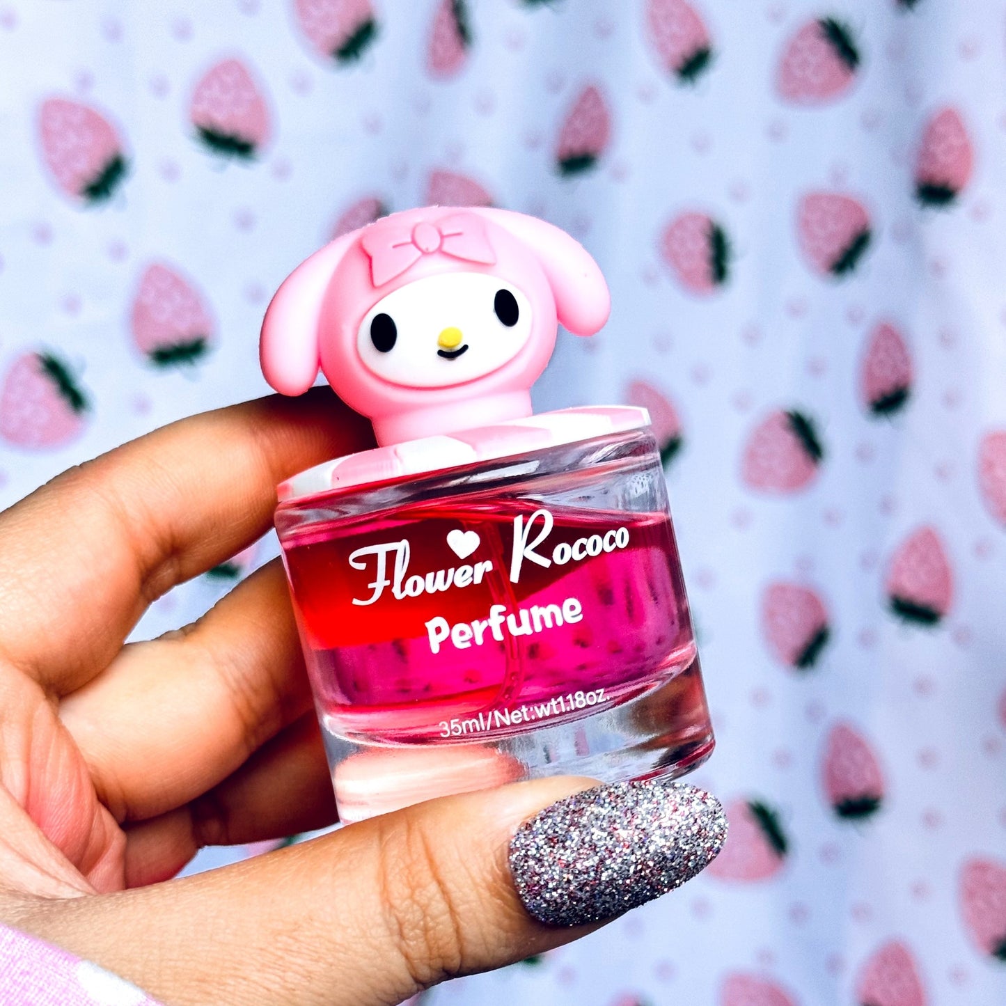 A hand with glittery silver nail polish holds a small perfume bottle shaped like a pink cartoon character. The bottle has pink liquid and a clear glass base, with the label reading 'Flower Rococo Perfume'.