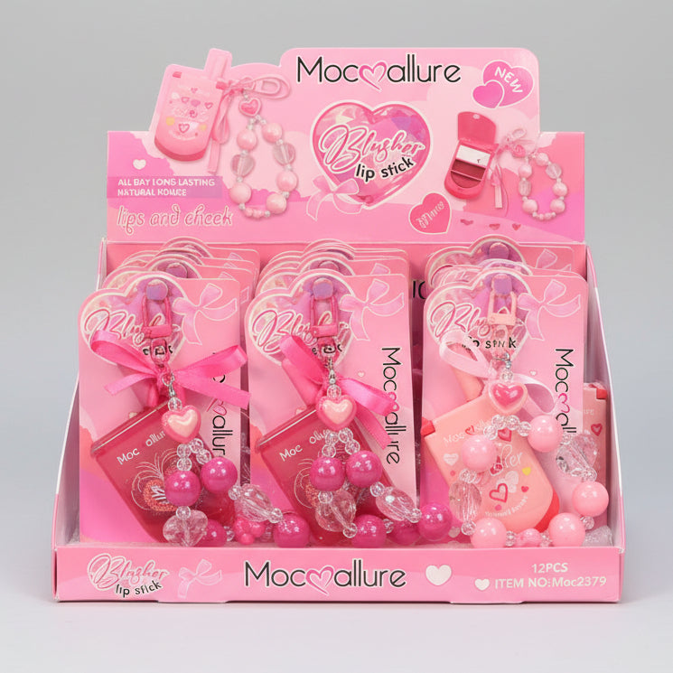A retail display box filled with pink Moccolure 'Blister Lip Stick' products, including small pink lip balm tubes, heart-shaped charms, and beaded keychains, photographed in a store setting.
