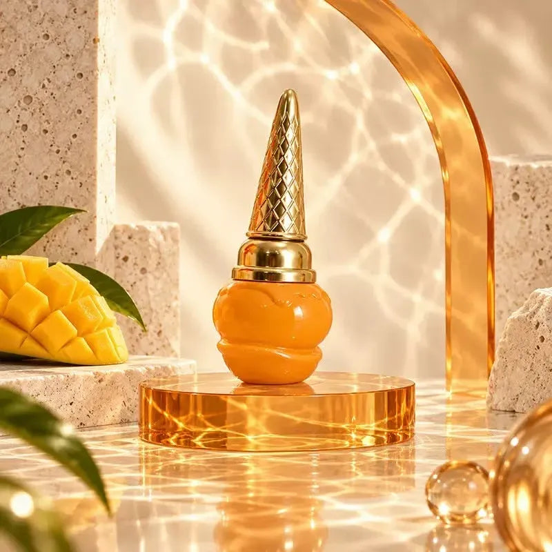 An orange and gold decorative bottle or perfume bottle displayed on a round gold pedestal, with warm sunlight and shadows across a stone-textured surface. Mango slices and leaves are partially visible at the left edge, and a translucent gold arch decorates the background.