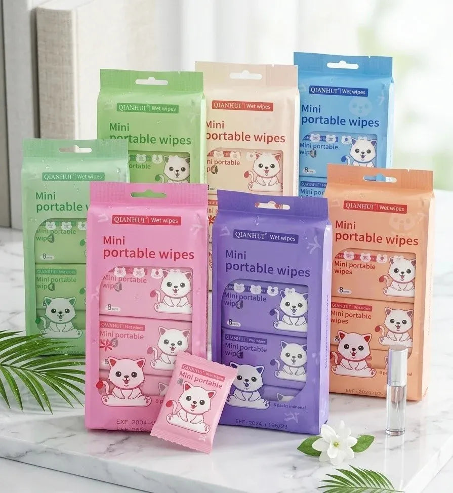 Six packs of QuanHui Mini Portable Wipes in assorted pastel colors (green, orange, blue, pink, purple) arranged on a white marble surface with decorative green leaves and a small white flower. A small clear spray bottle is visible on the right.