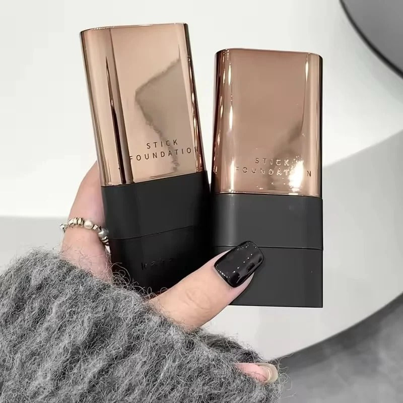 Two rectangular foundation sticks with metallic rose-gold caps and black bases are held in a hand wearing a gray fuzzy sweater and a silver ring. The embossed text on the caps reads 'STICK FOUNDATION'.