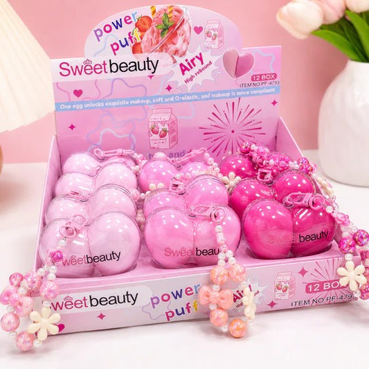 A pink retail display box filled with spherical cosmetic egg-shaped products branded Sweetbeauty, including pale pink, light pink, and bright pink eggs with pearl bead necklaces attached. The display card reads 'Sweetbeauty Power Puff Airy' and shows a 12-box pack.