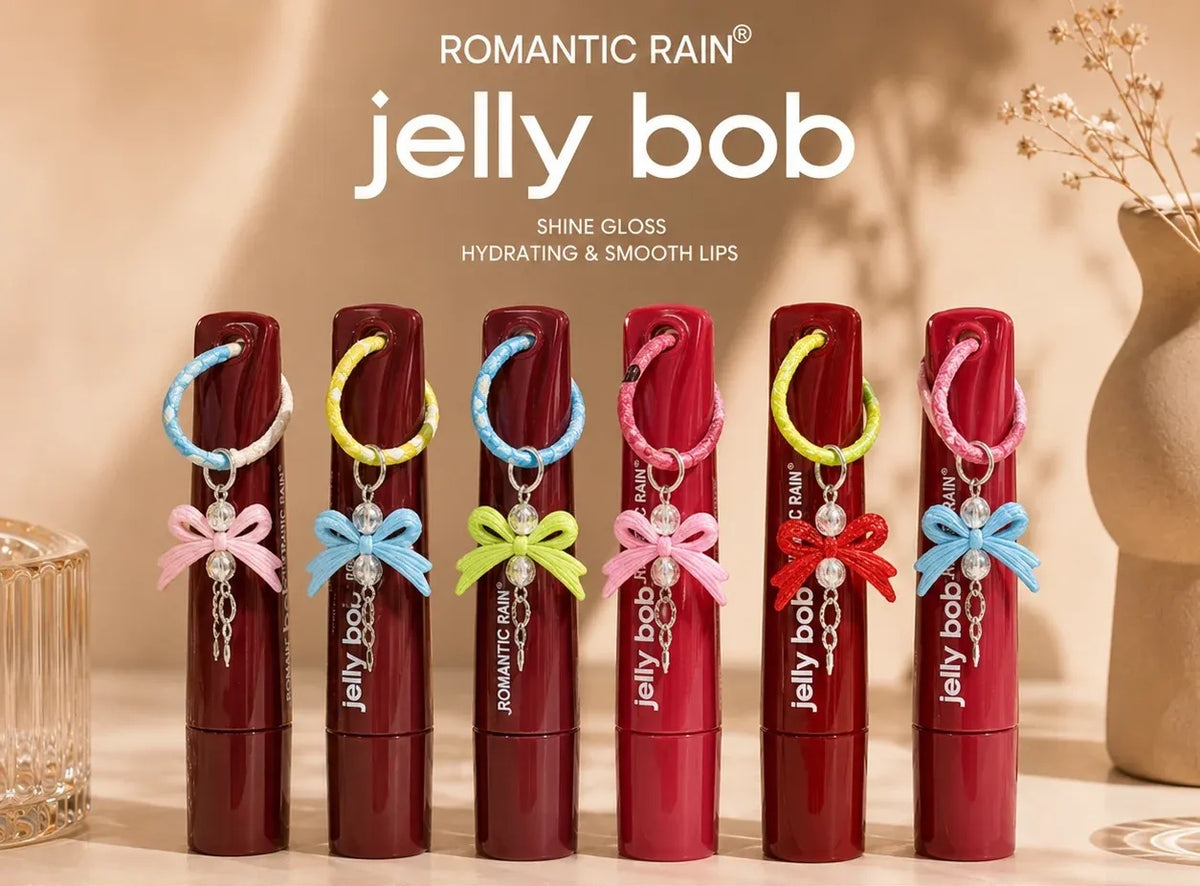 Six tubes of Jelly Bob shine gloss lip balm by Romantic Rain are lined up on a beige surface. Each tube has a red cap and a dark berry-red body, and they are decorated with colorful ribbon bows (pink, blue, yellow, pink, red, and blue) and silver chain charms.