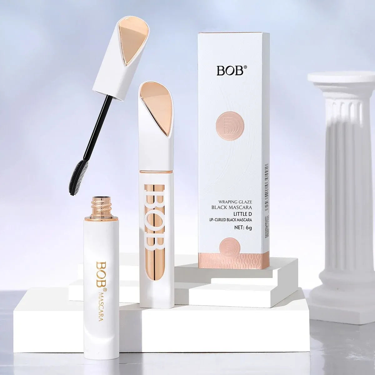 BOB Eyebrow Glaze Black Mascara shown with the open tube, wand, and retail box on white display blocks. The packaging is white with rose-gold/copper-toned accents and black text, photographed against a soft light blue studio background with a white column prop.