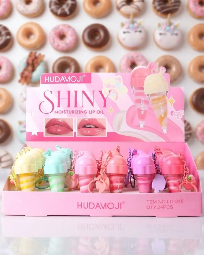 Hudamoji Shiny Moisturizing Lip Oil in a pink display box with multiple pastel lip oil tubes shaped like donuts, set against a background of decorated donuts.