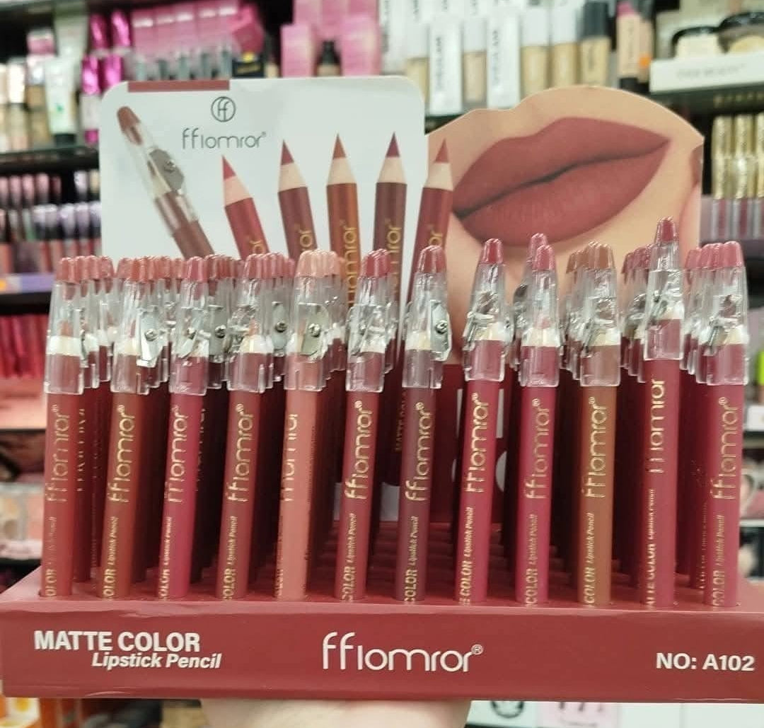 A display of fflomror matte color lipstick pencils in a pink and nude shade range, held by a hand with swatches on the wrist, set against a busy cosmetic store background