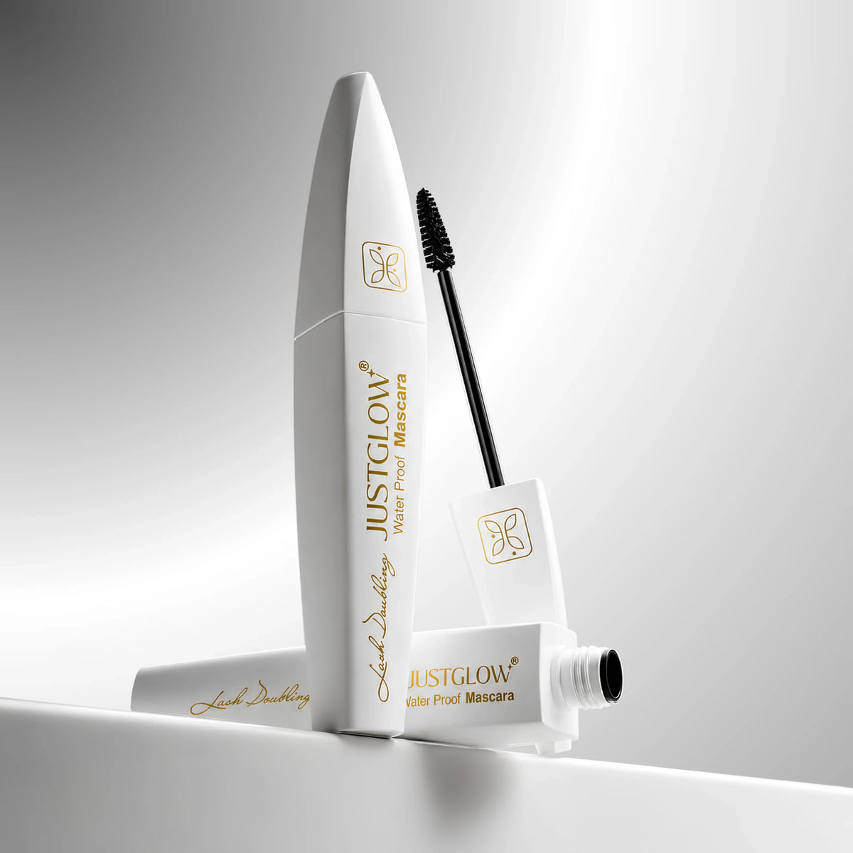 A white tube of JustGlow Water Proof Mascara with its applicator wand and cap, arranged on a reflective white surface against a soft gray gradient background.