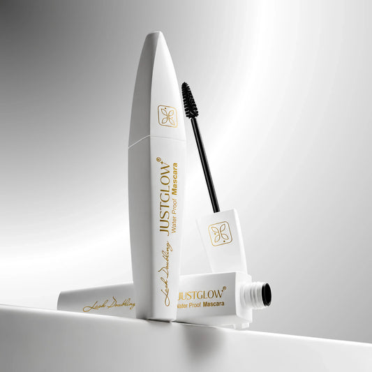 A white tube of JustGlow Water Proof Mascara with its applicator wand and cap, arranged on a reflective white surface against a soft gray gradient background.