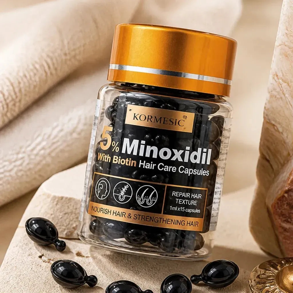 A bottle of KORMESIC 5% Minoxidil hair care capsules with a metallic gold cap, set on a textured beige surface with capsules scattered around.